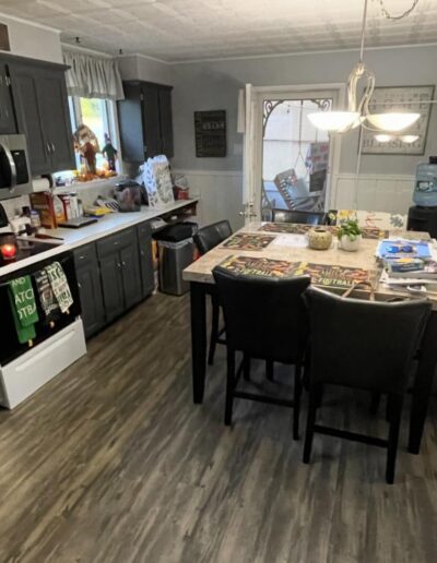 A kitchen with gray cabinets, stainless steel appliances, a small island with bar stools, and various items on the countertop. A door leads outside, and autumn decor is on the wall.
