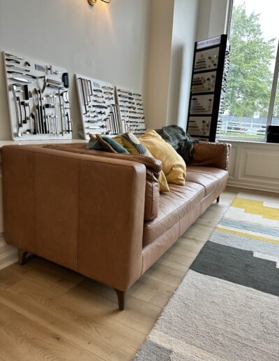 A tan leather couch with colorful pillows is in a well-lit room. Knives are displayed on wall racks in the background. A window shows a view of greenery outside.