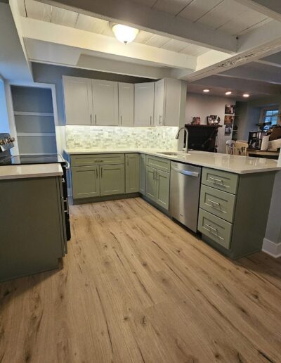 Modern kitchen with light wood flooring, green and white cabinets, stainless steel appliances, and a tile backsplash. Ceiling features white beams and recessed lighting.